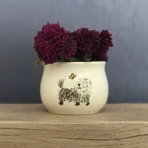 May include: White ceramic planter with a hand-painted illustration of a dog and a butterfly. The planter is filled with purple flowers.