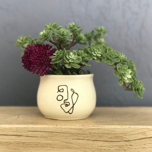 May include: A white ceramic pot with a black line drawing of a face on it. The pot is filled with green succulents and a single purple flower.