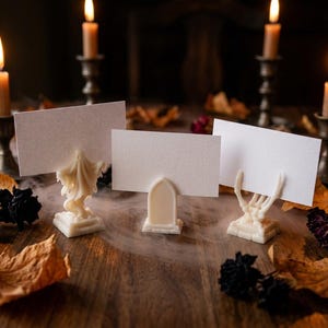 May include: Three white place card holders featuring a ghost, a tombstone, and a skeleton hand, each holding a blank rectangular white card. The holders are arranged in a row, with lit candles in metal holders and scattered dried leaves nearby.