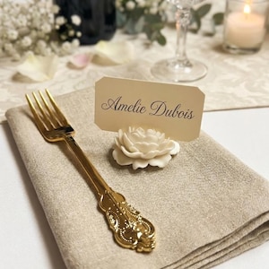 May include: A gold-toned metal fork with an ornate handle rests on a folded beige linen napkin next to a white flower-shaped place card holder. The holder supports a rectangular cream-colored card with the name Amelie Dubois printed in black.
