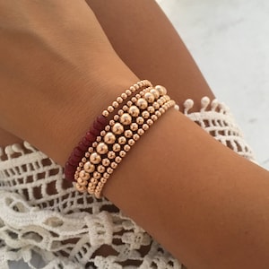 May include: Close-up of three gold beaded bracelets and one red beaded bracelet. The bracelets are worn on the wrist. The gold beads vary in size, creating a layered effect. The bracelets are on a tanned arm.