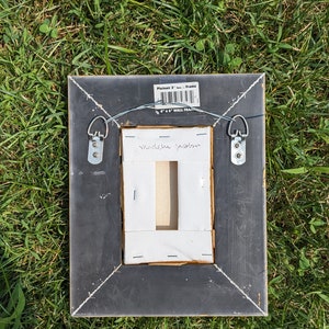 May include: Black picture frame with gold trim, featuring a white mount and a white label with the text "madeleine garden". The frame has two metal hanging hooks on the back and a label that reads "Plainair 3" Frame 4 x 6 WALL FRAME".