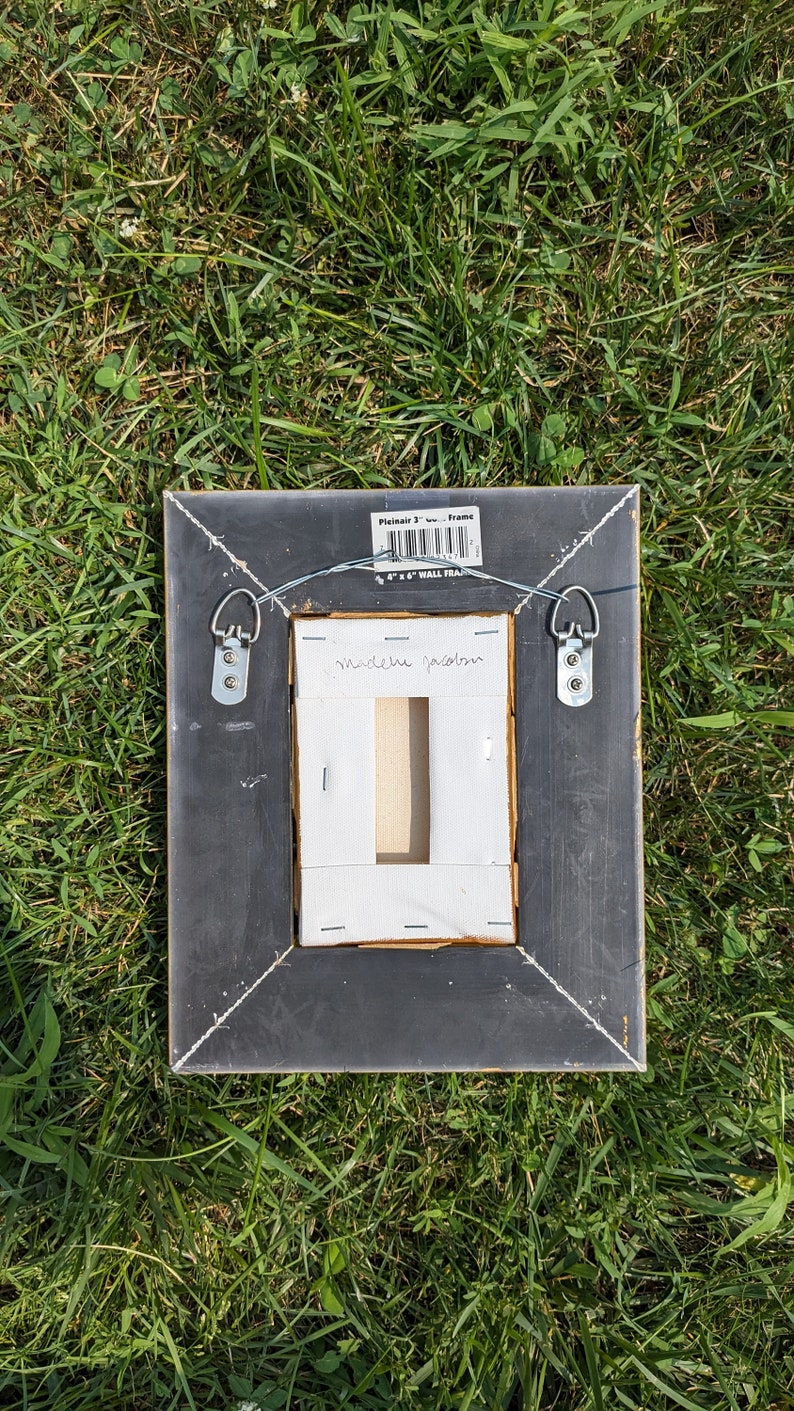 May include: Black picture frame with gold trim, featuring a white mount and a white label with the text "madeleine garden". The frame has two metal hanging hooks on the back and a label that reads "Plainair 3" Frame 4 x 6 WALL FRAME".