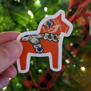 May include: A red and white Dala horse sticker with blue and yellow floral patterns. The sticker is on a green Christmas tree background.