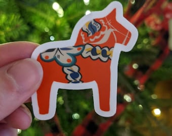 May include: A red and white Dala horse sticker with blue and yellow floral patterns. The sticker is on a green Christmas tree background.