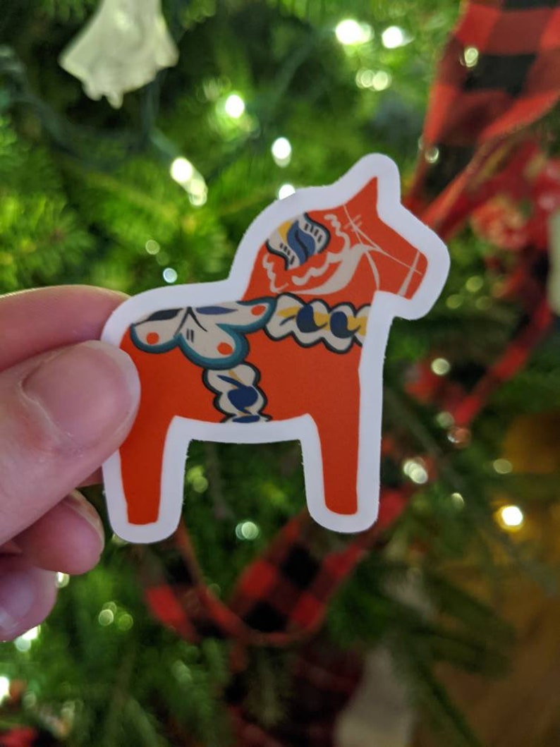 May include: A red and white Dala horse sticker with blue and yellow floral patterns. The sticker is on a green Christmas tree background.