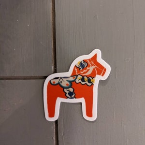 May include: A red and blue Dala horse sticker with white outlines. The horse is a traditional Swedish folk art design.