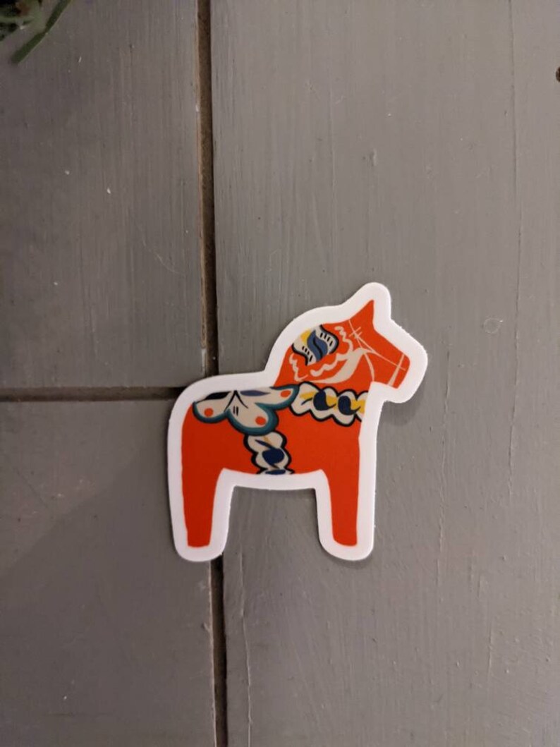 May include: A red and blue Dala horse sticker with white outlines. The horse is a traditional Swedish folk art design.