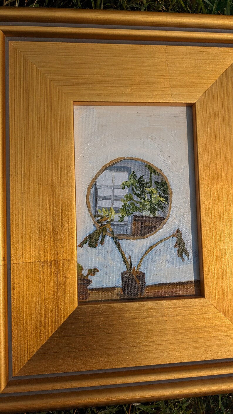 May include: A framed painting of a plant with a circular window reflection in the background. The painting is in a gold frame with a white mount.
