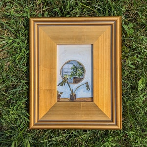 May include: A framed painting depicting a window with a plant in front of it. The window is round and the plant is green. The frame is gold.