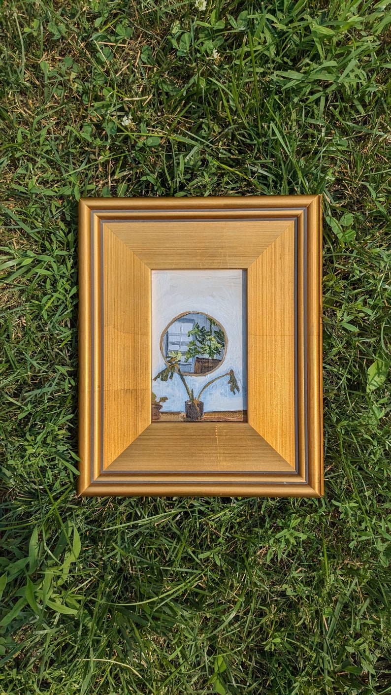 May include: A framed painting depicting a window with a plant in front of it. The window is round and the plant is green. The frame is gold.