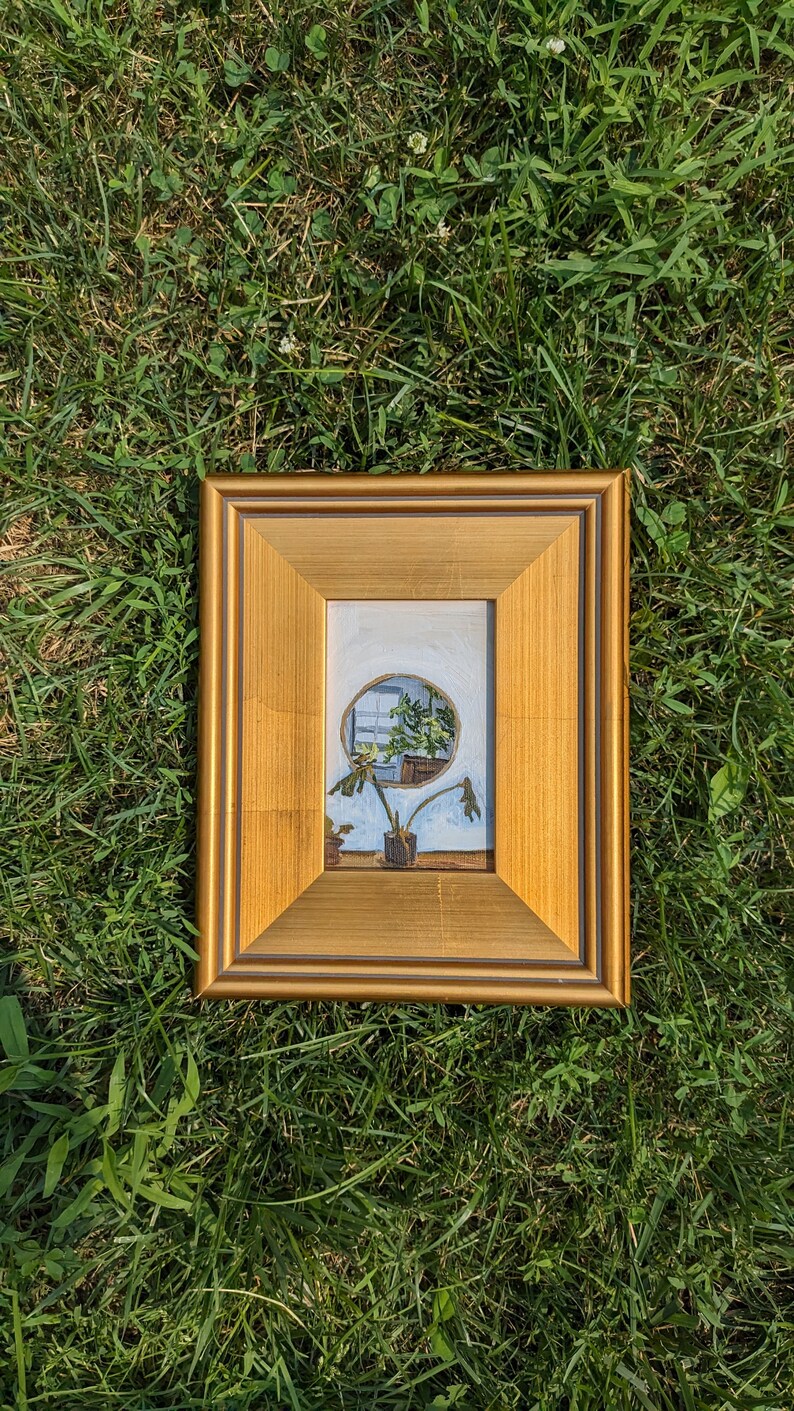 May include: A framed painting of a plant in a pot, viewed through a round window. The painting is in a gold frame and is set against a green grassy background.