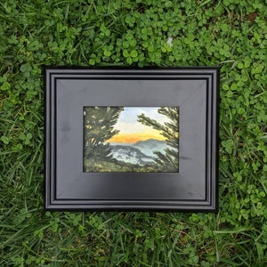 May include: A framed painting depicting a landscape scene with a sunset over a mountain range. The painting is in a black frame and is resting on a bed of green grass.