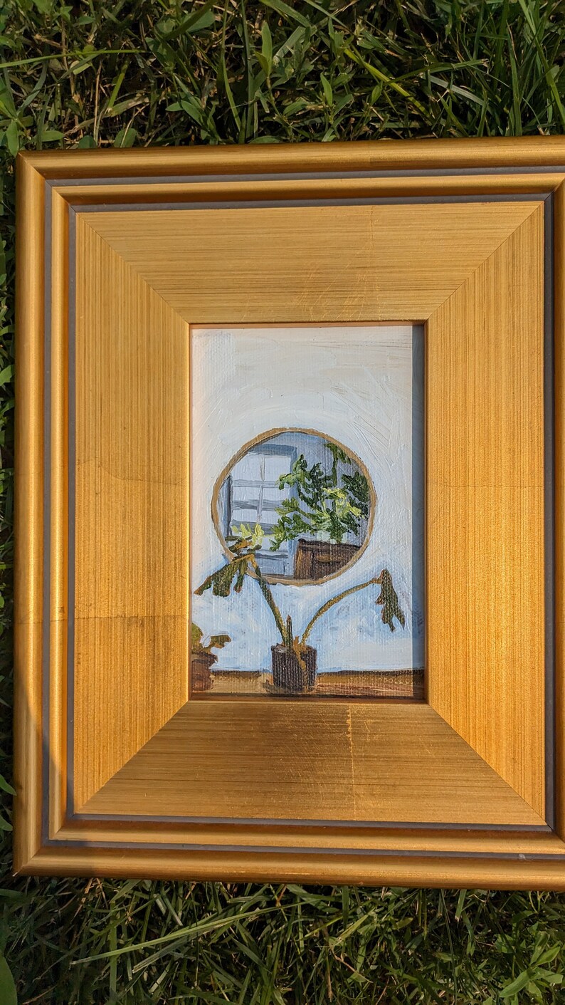 May include: A framed painting of a plant in a pot, with a circular window behind it showing a view of another plant. The painting is in a gold frame with a dark grey inner border.
