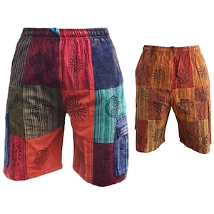 Puede incluir: Dos pantalones cortos patchwork. Un par presenta una mezcla de paneles rojos, naranjas, azules y verdes con diseños de sol y geométricos negros. El segundo par es naranja y marrón con diseños negros similares. Ambos tienen cinturillas elásticas.