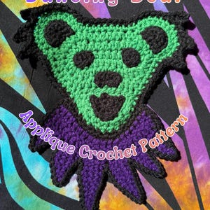 May include: A handmade crochet applique in the shape of a bear with a heart-shaped head. The bear features a green face, black eyes and nose, and a purple body. The text "Dancing Bear" and "Applique Crochet Pattern" are written on the image.