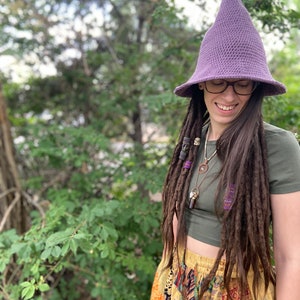 May include: A person wearing a purple crochet witch hat with a wide brim and a green shirt. They have long brown dreadlocks with beads and are wearing a colorful patchwork skirt.