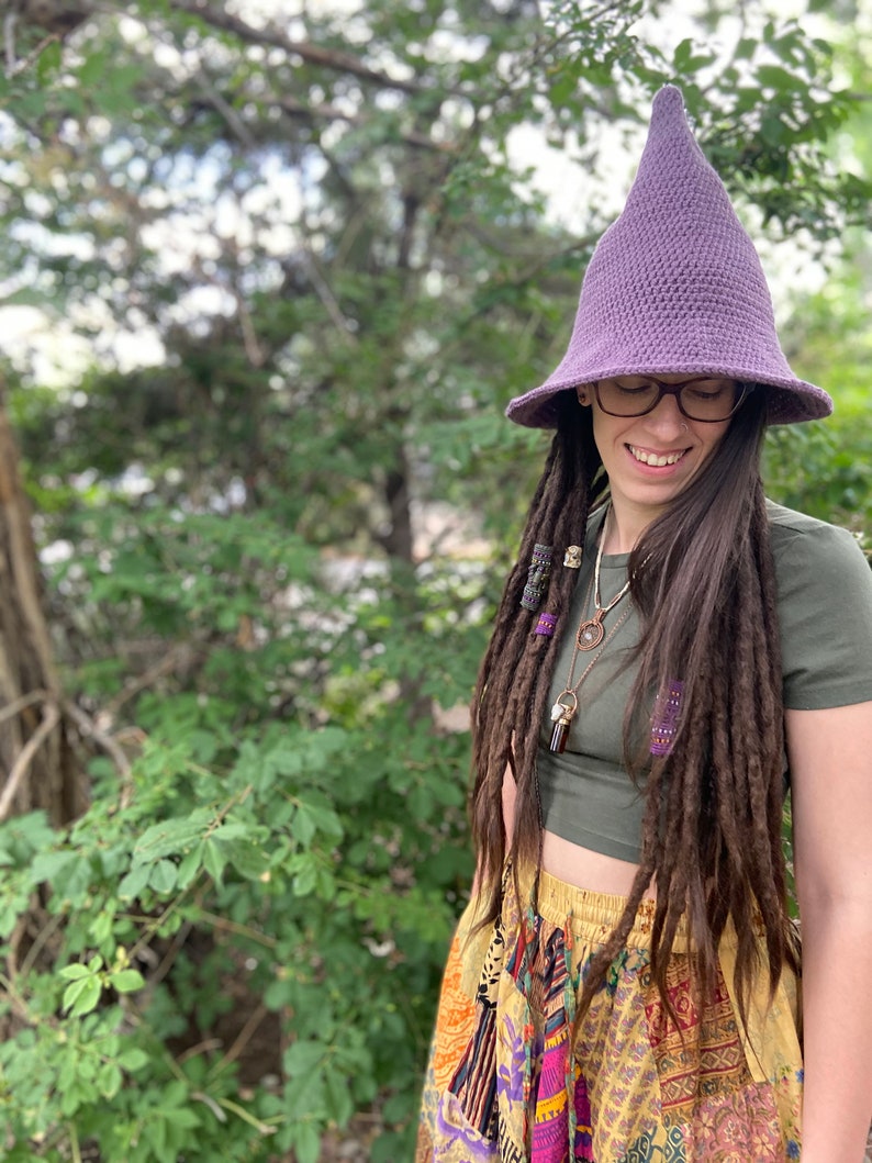 May include: A person wearing a purple crochet witch hat with a wide brim and a green shirt. They have long brown dreadlocks with beads and are wearing a colorful patchwork skirt.