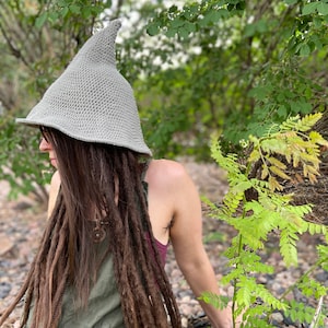 May include: A person wearing a gray crocheted pointed hat with a long, brown braid. The person is sitting in a forest setting with green foliage.