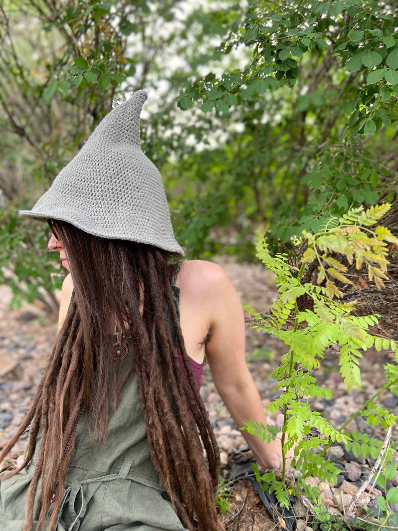 May include: A person wearing a gray crocheted pointed hat with a long, brown braid. The person is sitting in a forest setting with green foliage.