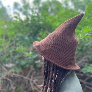 May include: A brown crocheted witch hat with a pointed tip. The hat is worn by a person with long brown dreadlocks.
