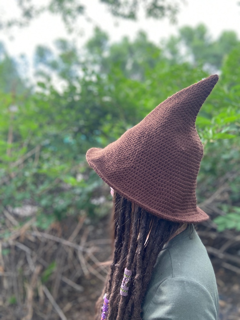 May include: A brown crocheted witch hat with a pointed tip. The hat is worn by a person with long brown dreadlocks.