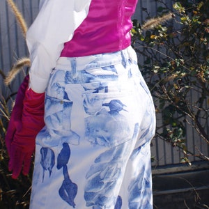 May include: A woman wearing a white button-down shirt, a pink satin corset, and white jeans with a blue floral pattern. She is wearing a pink headband and has her hair pulled back. She is standing in front of a metal wall with a bush of pink flowers in the background.
