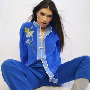 May include: A woman wearing a blue silk pajama set with a floral embroidery design. The set includes a long-sleeved shirt with a mandarin collar and matching pants. She is wearing gray boots.