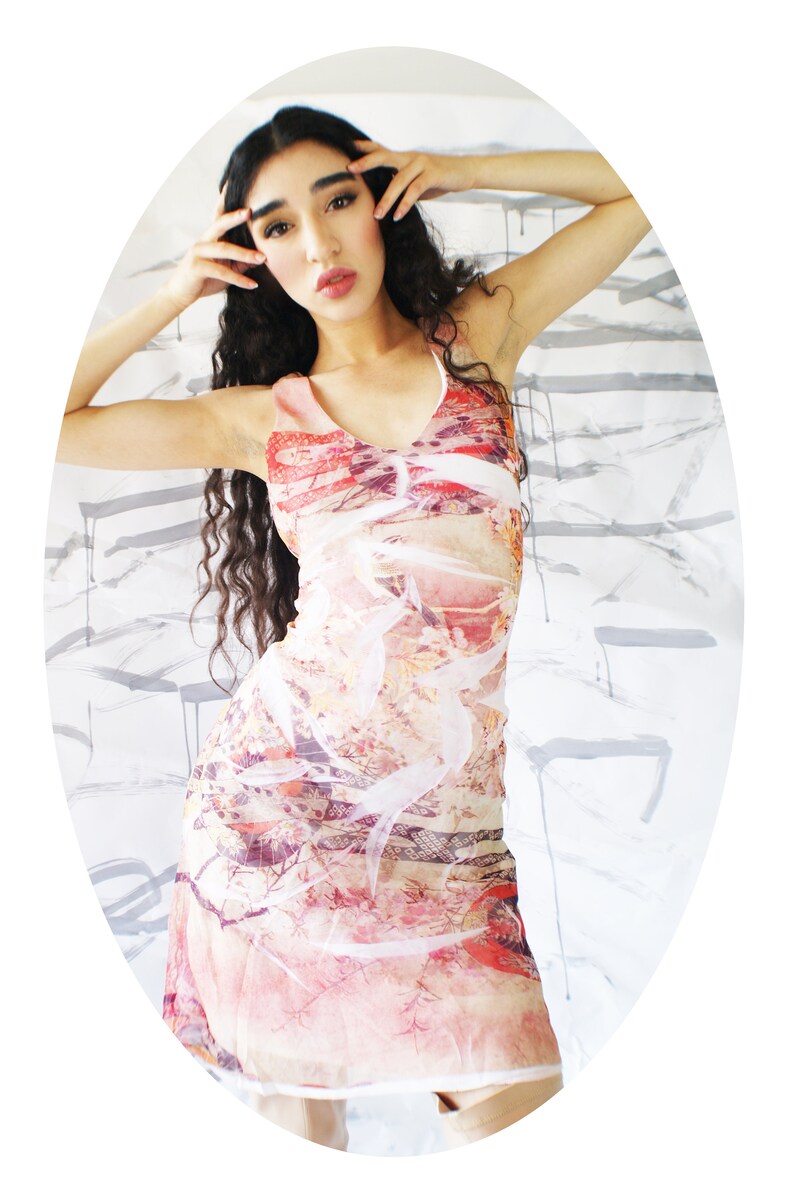 Puede incluir: Una mujer lleva un vestido sin mangas con estampado floral de colores. El vestido tiene un escote en V y una silueta ajustada. El fondo es una pared blanca con salpicaduras de pintura gris.