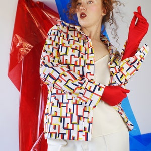 May include: A woman wearing a white shirt with a geometric pattern in red, yellow, and blue. She is wearing white pants and red gloves. The background is a red and blue plastic sheet.