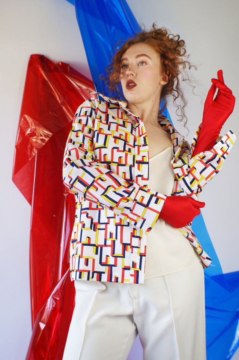 May include: A woman wearing a white shirt with a geometric pattern in red, yellow, and blue. She is wearing white pants and red gloves. The background is a red and blue plastic sheet.