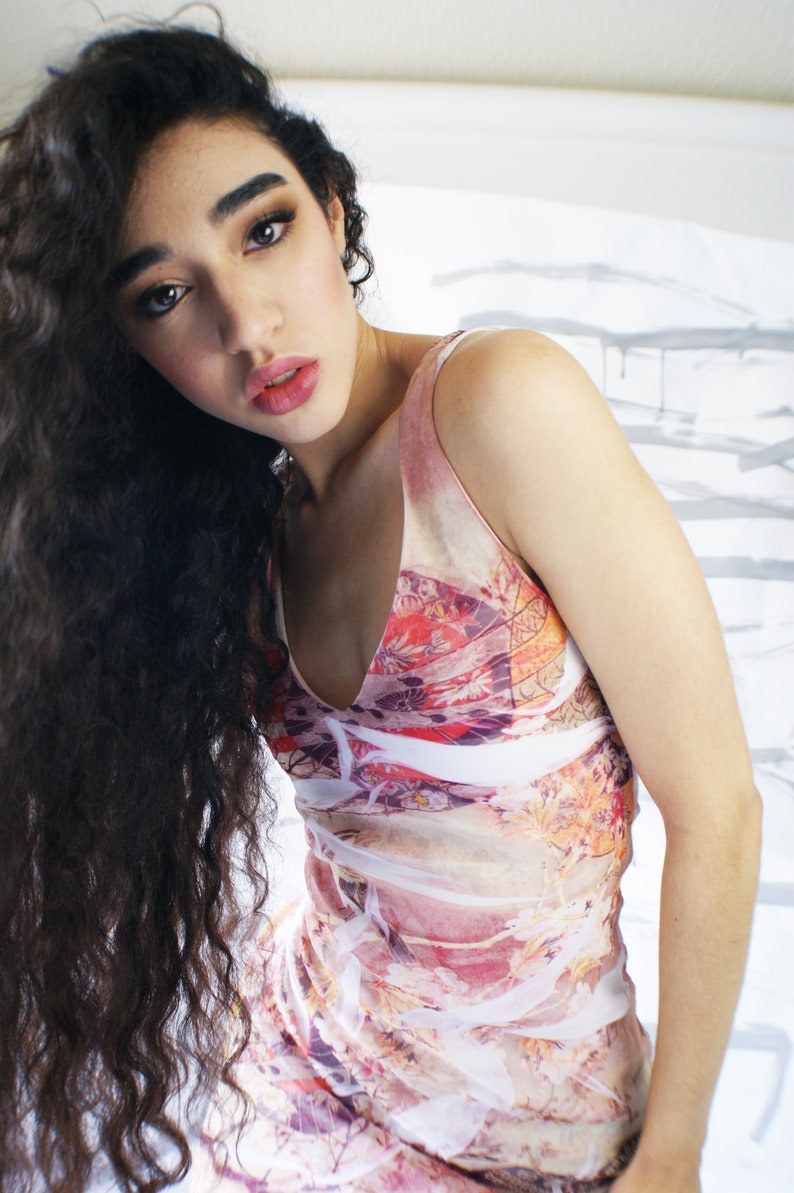 Puede incluir: Una mujer con el pelo largo y rizado oscuro lleva un vestido blanco sin mangas con estampado floral colorido. El vestido tiene un escote en V y un corte holgado y fluido.