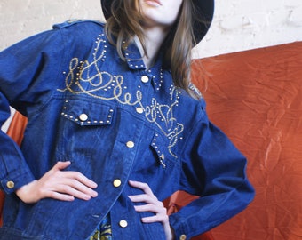 Peut inclure: Une femme portant une veste en jean bleue avec des broderies dorées et un chapeau noir. La veste a une fermeture à boutons et est décorée de clous dorés. La femme porte une jupe à motifs colorés.