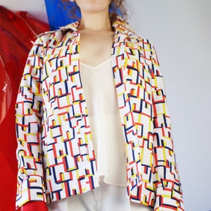 May include: A white button-up shirt with a geometric pattern in red, yellow, and blue. The shirt has long sleeves and a collar. The model is wearing white pants and red gloves.
