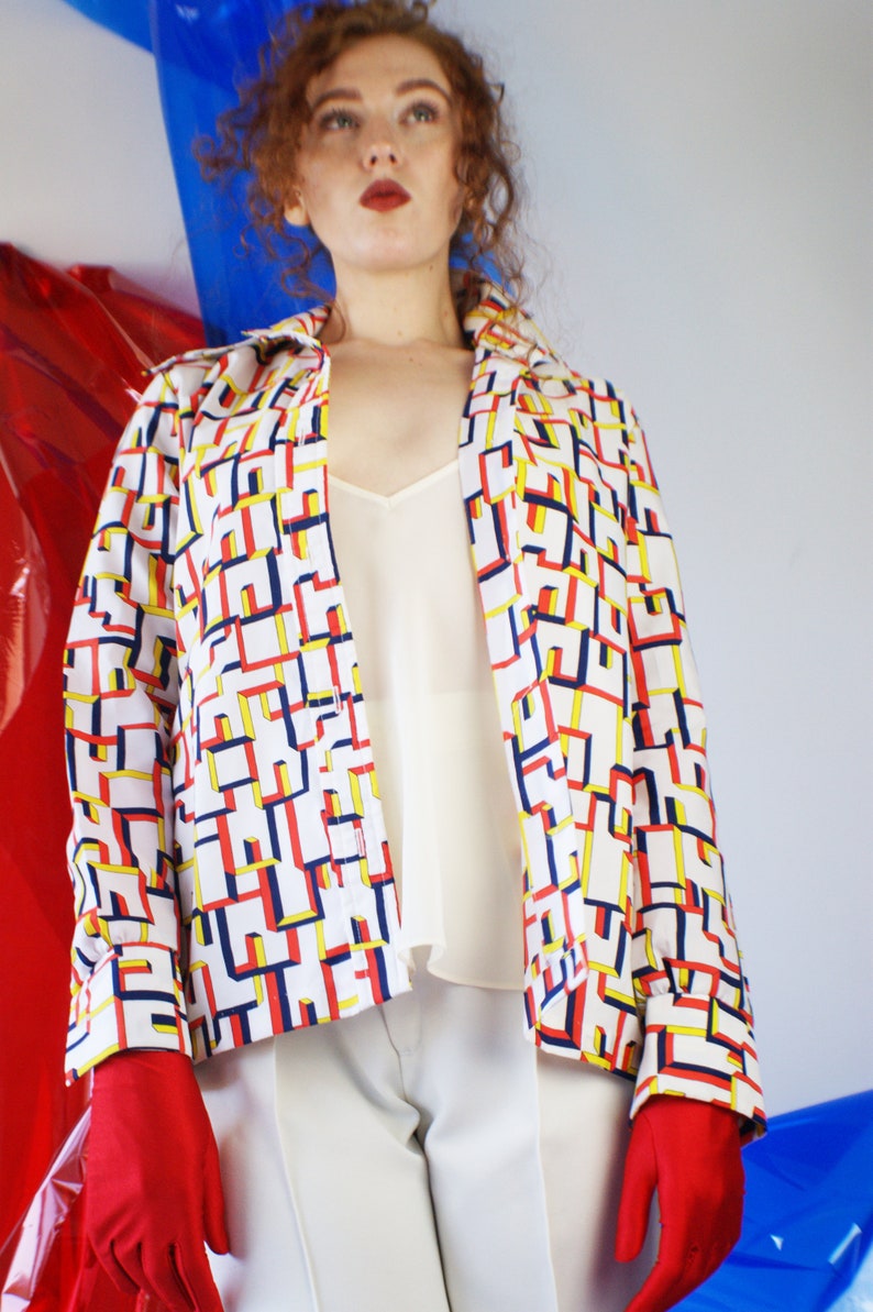 May include: A white button-up shirt with a geometric pattern in red, yellow, and blue. The shirt has long sleeves and a collar. The model is wearing white pants and red gloves.