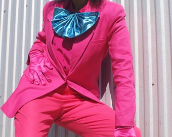 May include: A woman wearing a hot pink suit with a blue bow tie and matching gloves. She is standing in front of a silver corrugated metal wall.