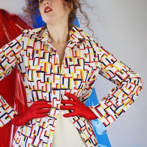 May include: A white shirt with a geometric pattern in red, yellow, and blue. The shirt has a collar and long sleeves. The model is wearing red gloves and white pants.