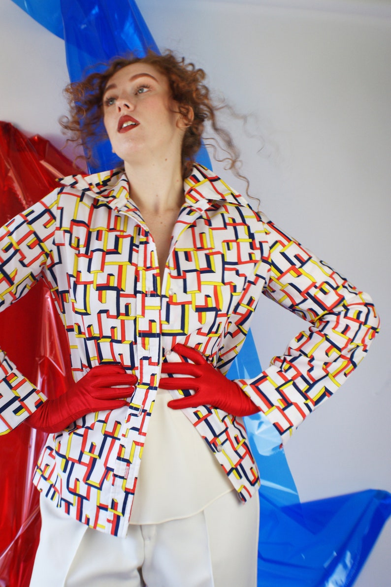 May include: A white shirt with a geometric pattern in red, yellow, and blue. The shirt has a collar and long sleeves. The model is wearing red gloves and white pants.