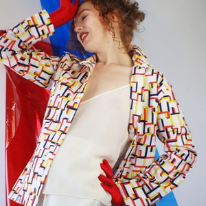 May include: A woman wearing a white top and white pants with a geometric patterned jacket in red, yellow, and blue. She is wearing red gloves and has curly red hair.