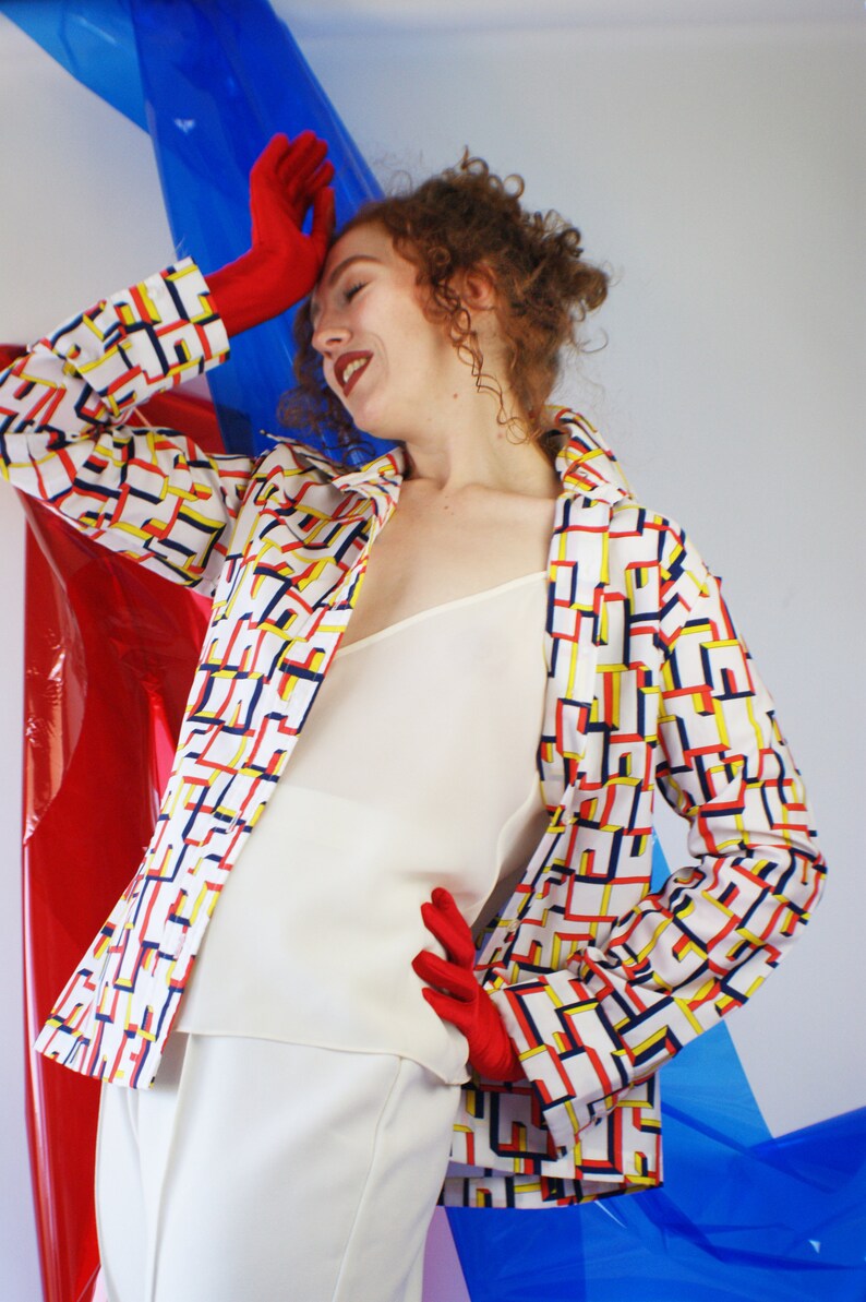 May include: A woman wearing a white top and white pants with a geometric patterned jacket in red, yellow, and blue. She is wearing red gloves and has curly red hair.