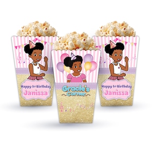 May include: Three pink and gold popcorn boxes with a musical theme. Each box features a cartoon illustration of a young girl wearing a pink dress and a pink bow in her hair. The boxes are decorated with musical notes, balloons, and the text "Happy 1st Birthday Janissa" and "Gracie's Corner".