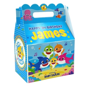 May include: A blue Baby Shark themed party favor box with a handle. The box features the text "HAPPY 3RD BIRTHDAY James" and illustrations of cartoon sharks, fish, and an octopus. The side of the box says "THANKS FOR COMING".