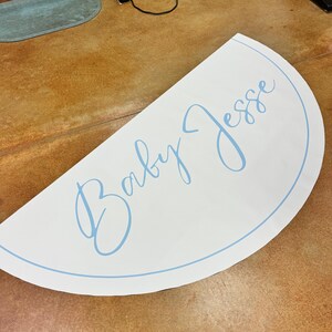 May include: A white, semi-circular sign with the words "Baby Jesse" in light blue script. The text is inside a light blue border. The sign is on a brown surface.