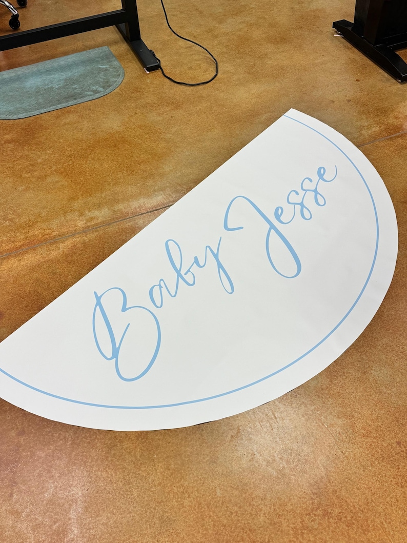 May include: A white, semi-circular sign with the words "Baby Jesse" in light blue script. The text is inside a light blue border. The sign is on a brown surface.