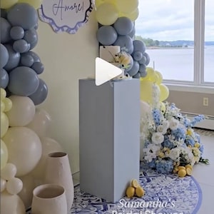 May include: A bridal shower scene with a cake on a gray pedestal, surrounded by yellow, white, and gray balloons. A floral arrangement with blue and white flowers and lemons is on the floor. The words "It's Amore!" are on a sign.