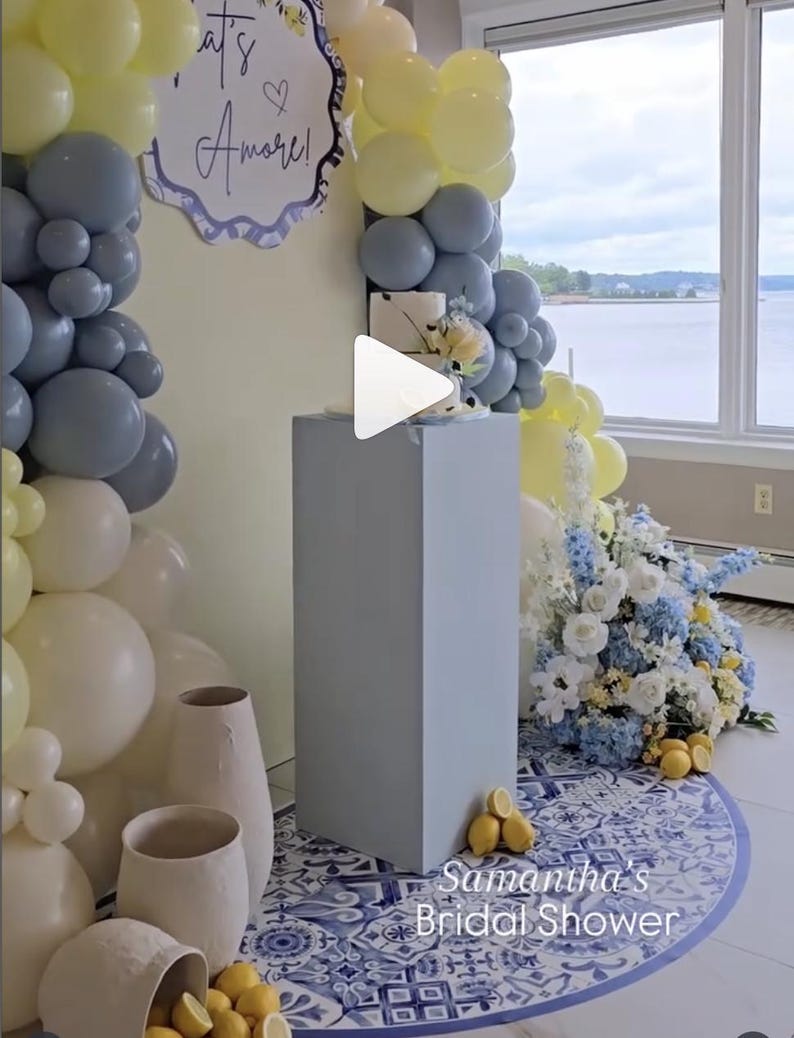 May include: A bridal shower scene with a cake on a gray pedestal, surrounded by yellow, white, and gray balloons. A floral arrangement with blue and white flowers and lemons is on the floor. The words "It's Amore!" are on a sign.