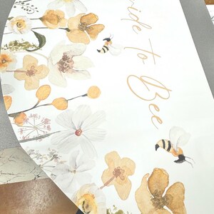 May include: A white, semi-circular sign with watercolor floral and bee illustrations. The design features yellow and white flowers, green foliage, and two bees. The words "Bride to Bee" are written in gold script.