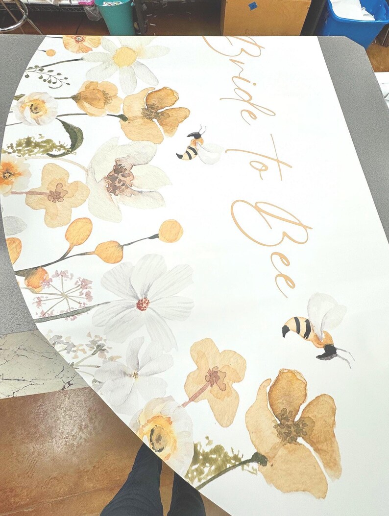 May include: A white, semi-circular sign with watercolor floral and bee illustrations. The design features yellow and white flowers, green foliage, and two bees. The words "Bride to Bee" are written in gold script.