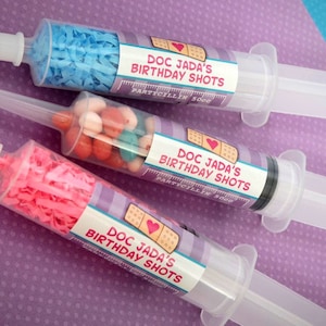 May include: Three clear plastic syringes decorated with colorful labels that say "Doc Jada's Birthday Shots" and "Partycillin 500g". The syringes are filled with blue and pink tissue paper and colorful candy.