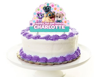 May include: A cake topper featuring characters from the animated series PAW Patrol. The topper is pink, blue, and green with paw prints and the text "Happy 2nd Birthday Charlotte".